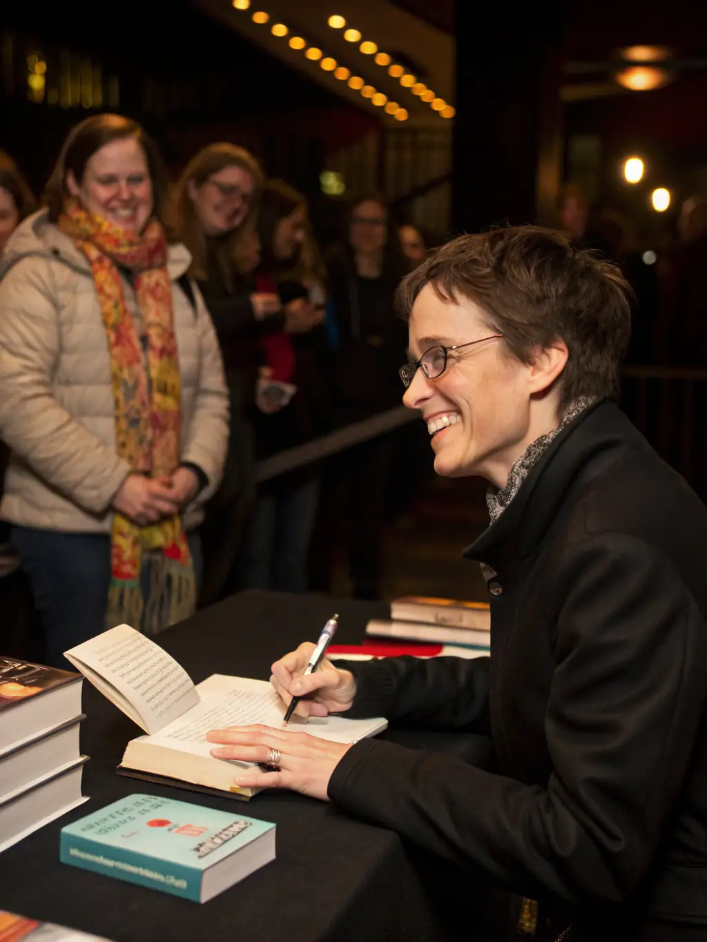 A photograph of a local author signing books at a CULTISSIME event, with a line of people waiting to meet the author, bookshelves in the background, and a banner with the CULTISSIME logo.